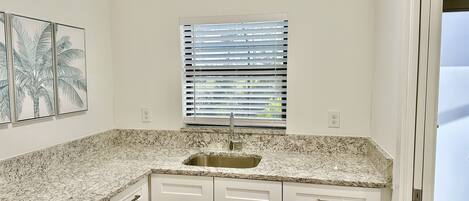 A kitchen with white cabinets, a granite countertop, a window with blinds, and a wall-mounted power outlet.