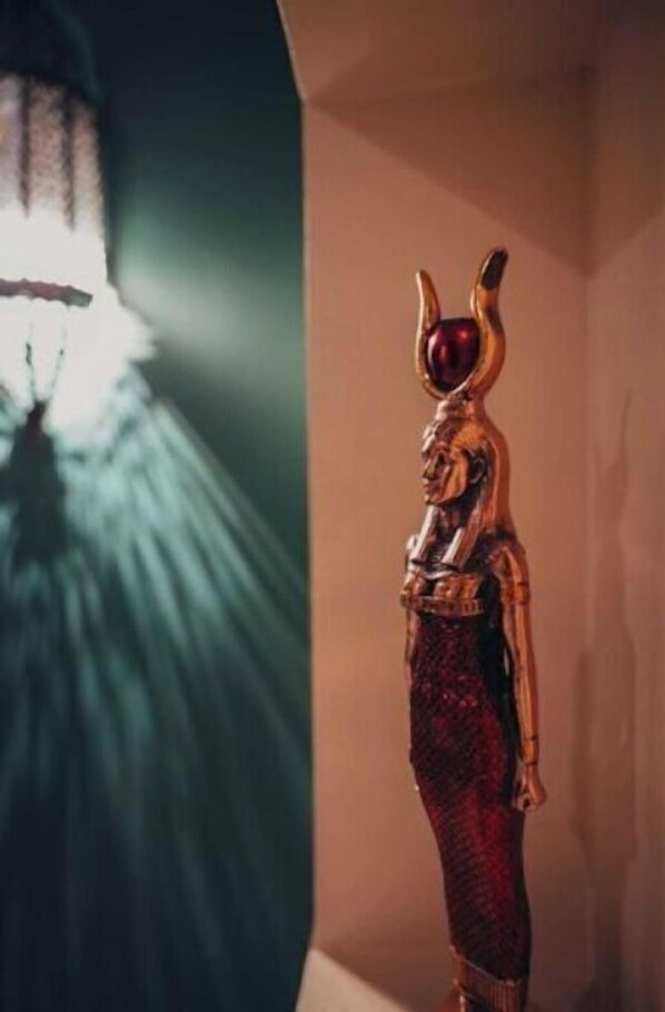 Eine Statue einer Figur mit Kopfschmuck und einem roten Juwel, stehend in einem Raum mit grüner Wand und einer Leuchte.