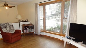 Una sala de estar con un sillón rojo, una estantería, un televisor y un ventanal con cortinas.