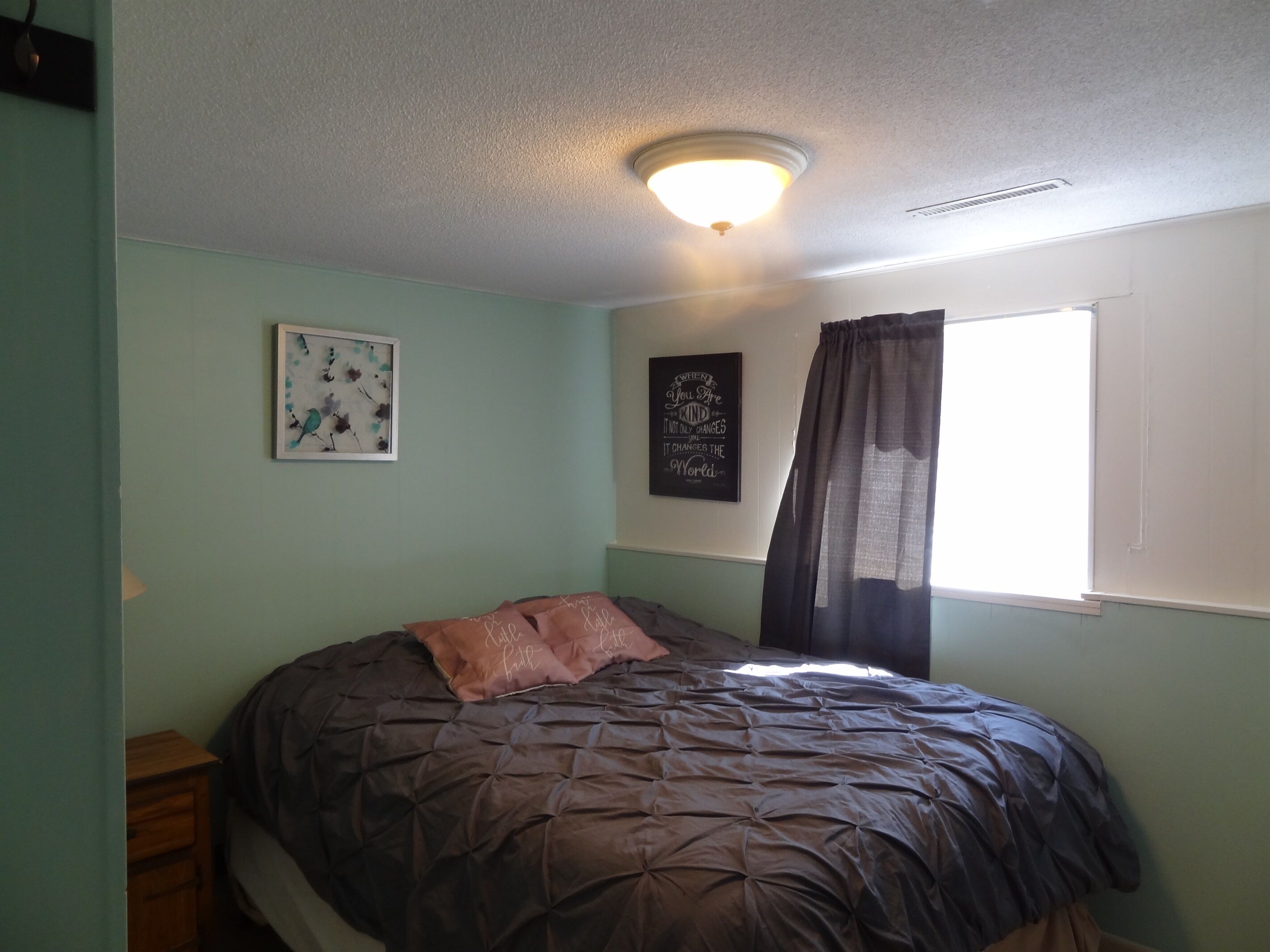 A bedroom with a bed, a nightstand, a wall-mounted light fixture, a window with curtains, and two framed pictures.