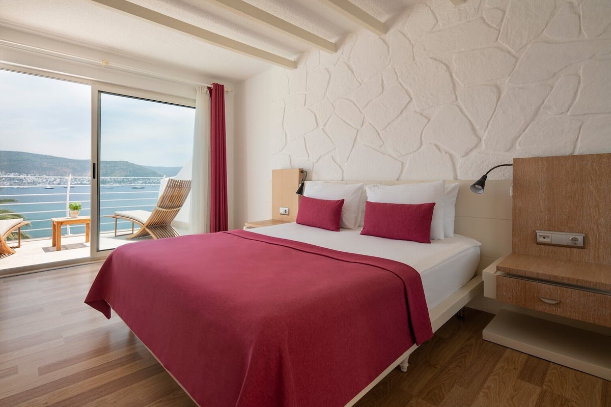 Une chambre d’hôtel avec un grand lit, une table de chevet en bois, un bureau et une chaise. La fenêtre offre une vue sur la mer et les collines.