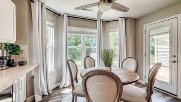 Un comedor con una mesa redonda, cuatro sillas, un ventilador de techo y ventanas grandes con cortinas.