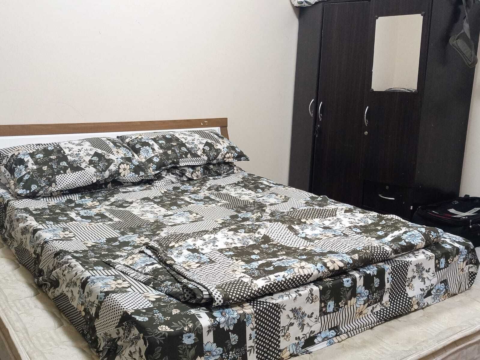 Een bed met een bloemenprint dekbed en kussens, een houten hoofdbord en een donkerkleurige garderobe met spiegel.