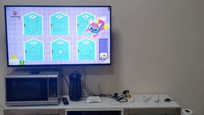 Televisão exibindo um jogo, um micro-ondas e um pequeno armário com um livro e uma garrafa de água.