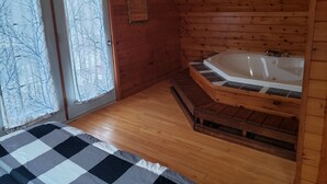 L’intérieur d’un chalet en bois, avec un lit à motifs à carreaux, un plancher en bois, une salle de bain dotée d’une baignoire et une porte en verre dépoli.