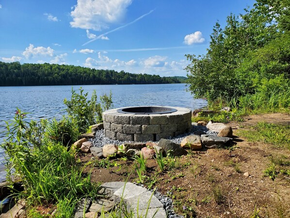A stone fire pit surrounded by rocks and vegetation near a lake. - Cozy cottage with private waterfront & wifi (Bancroft)