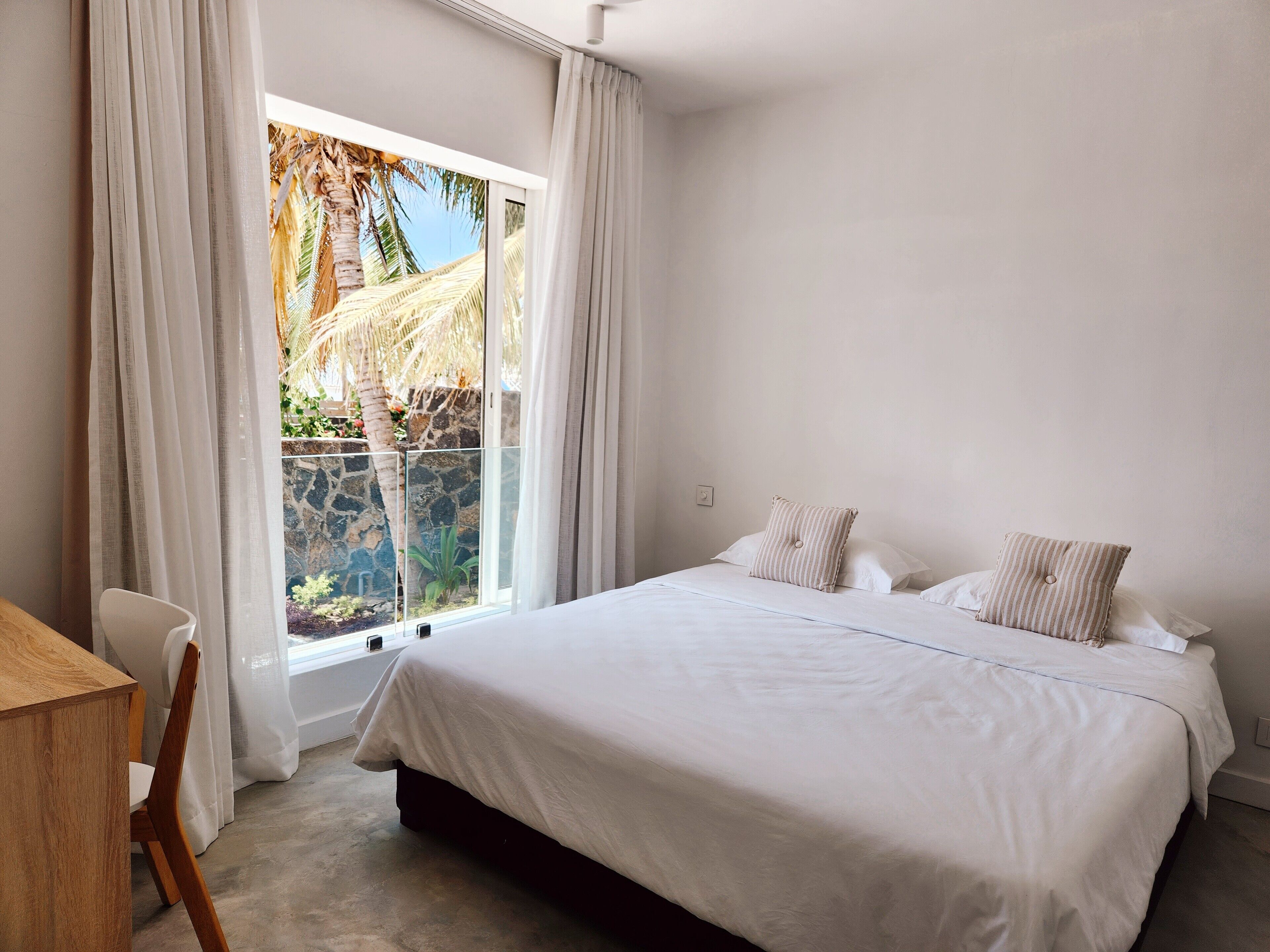 A bedroom with a large bed, white curtains, and a view of palm trees and a stone wall.