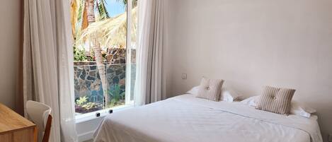 A bedroom with a large bed, white curtains, and a view of palm trees and a stone wall.