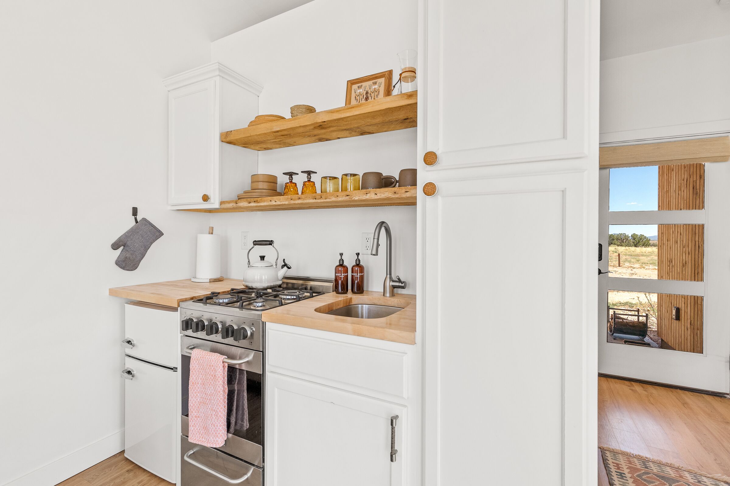 A kitchen with white cabinets, a wooden countertop, a stove, and a sink. There are shelves above the counter holding jars and a framed picture.