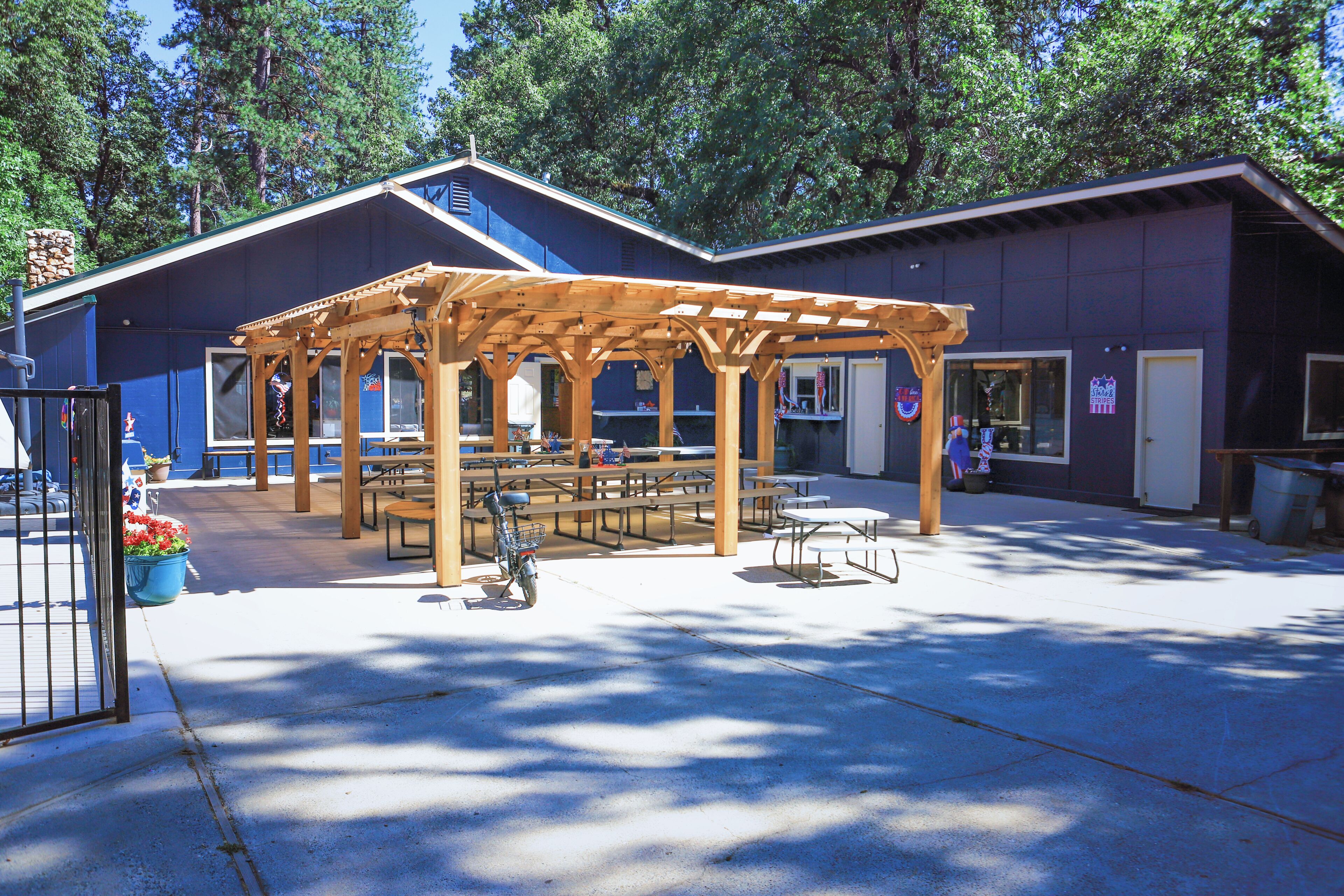 An outdoor covered picnic area with tables and benches, a wooden pergola, and a building with windows and a door in the background.