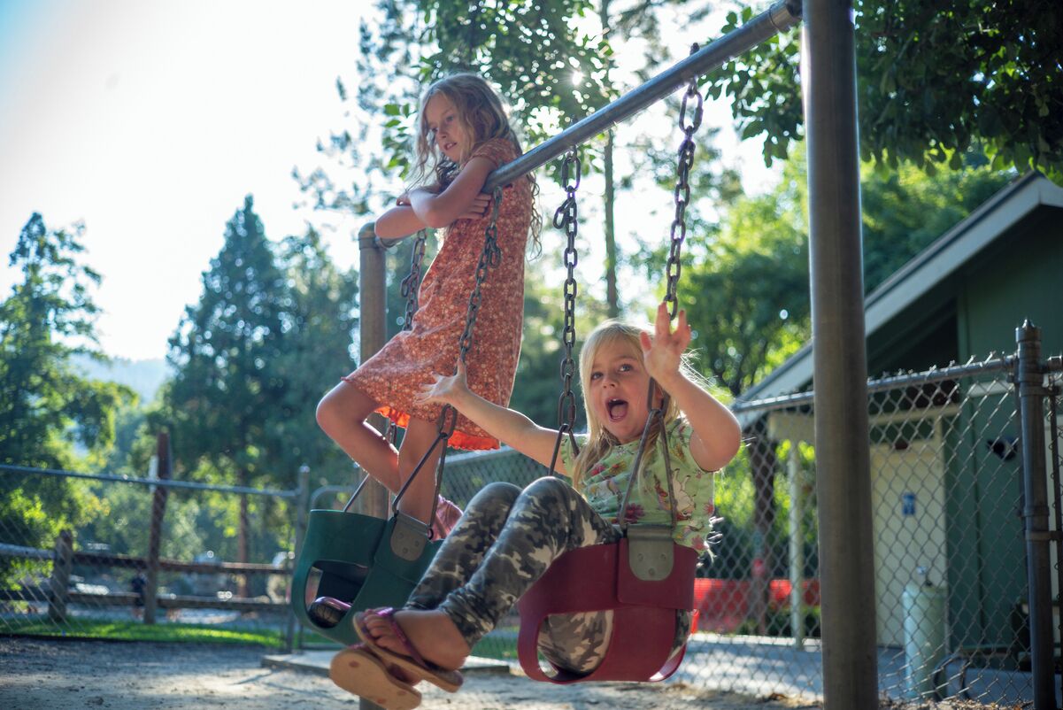 Two children on swings in a fenced outdoor area with trees and a building in the background.