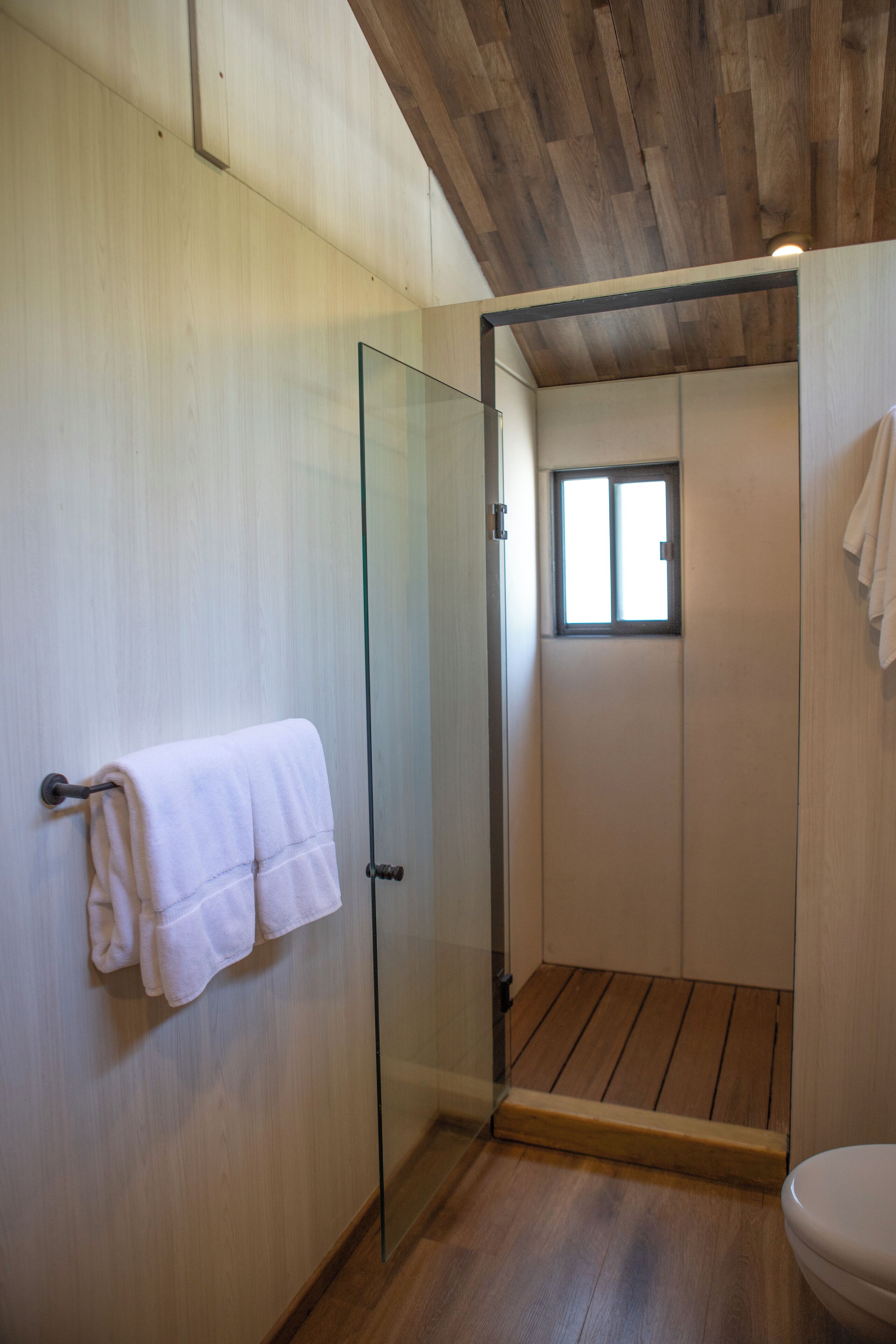 A bathroom with a glass shower door, wooden flooring, and a white towel hanging on the wall.