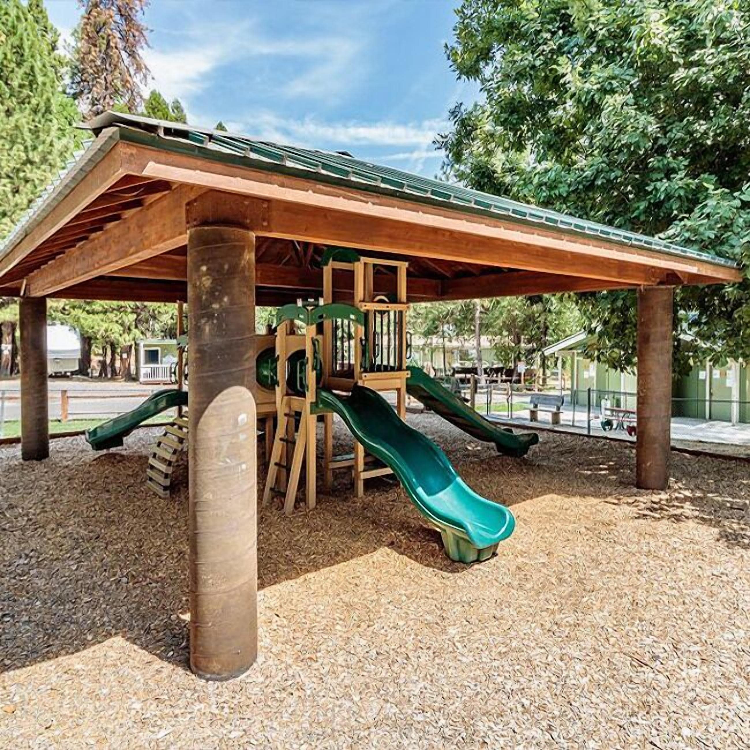 A wooden play structure with two slides and a climbing area, set on a gravel surface with trees in the background.