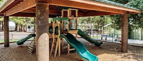 A wooden play structure with two slides and a climbing area, set on a gravel surface with trees in the background.