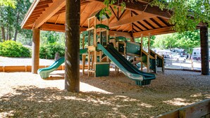 Une structure de jeux en bois, dotée de toboggans et d’éléments d’escalade, entourée d’arbres et recouverte d’un toit en bois.