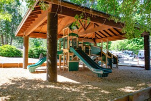 Una estructura de juego de madera con toboganes y elementos para escalar, rodeada de árboles y cubierta por un techo de madera.