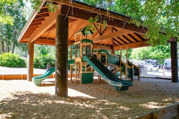 Eine Holzspielkonstruktion mit Rutschen und Kletterelementen, umgeben von Bäumen und mit einem Holzdach bedeckt.