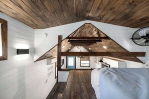 A bedroom with a sloped ceiling, wooden beams, a bed, a fan, and a view to an adjacent room. - Life's a Beach - 3 Acres with Hot Tub (Fredericksburg)