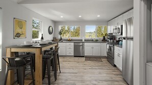 A modern kitchen with white cabinetry, stainless steel appliances, and a wooden dining table with bar stools. - Forest Heights ~ Relaxed Modern-Boho Style, Forest Views, Hot Tub, Wine Country (Forestville)