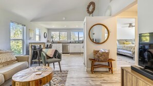 A spacious living room with a sofa, a coffee table, and a television. There is a kitchen area with a sink and cabinets, and a dining area with chairs. - Forest Heights ~ Relaxed Modern-Boho Style, Forest Views, Hot Tub, Wine Country (Forestville)