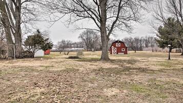 Un paysage rural avec une grange rouge, un grand arbre et un petit bâtiment blanc en arrière-plan.