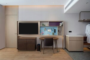 Una habitación moderna y compacta con televisión de pantalla plana, un escritorio con silla, un mueble pequeño y un estante empotrado con varios objetos.