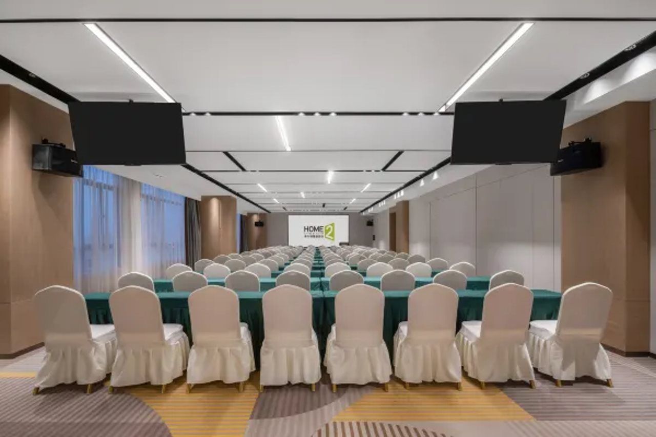 Une salle de conférence avec des rangées de tables et de chaises, un grand écran à l’avant et une pancarte indiquant « HOME 2 ».