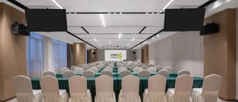 A conference room with rows of tables and chairs, a large screen at the front, and a sign reading 'HOME 2'.
