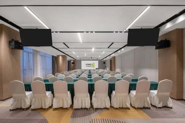 Un salón de conferencias con filas de mesas y sillas, una pantalla grande al frente y un letrero que dice 'HOME 2'.