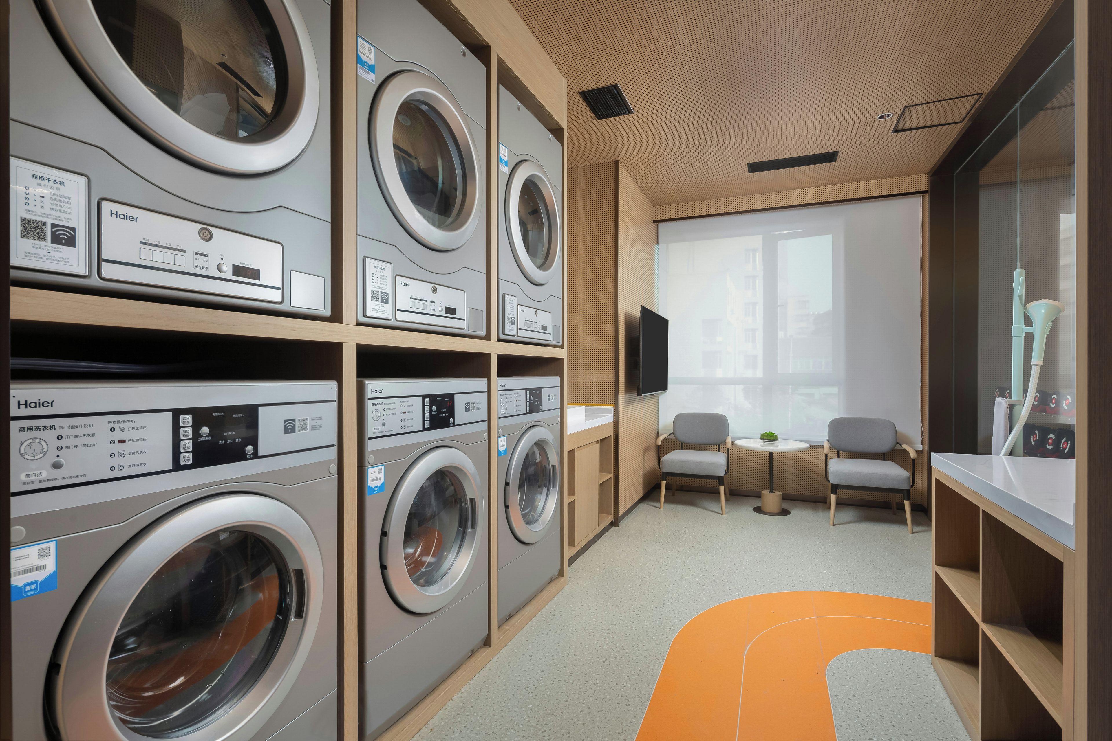 A laundry room with multiple Haier washing machines, a seating area with chairs, and a large window providing natural light.