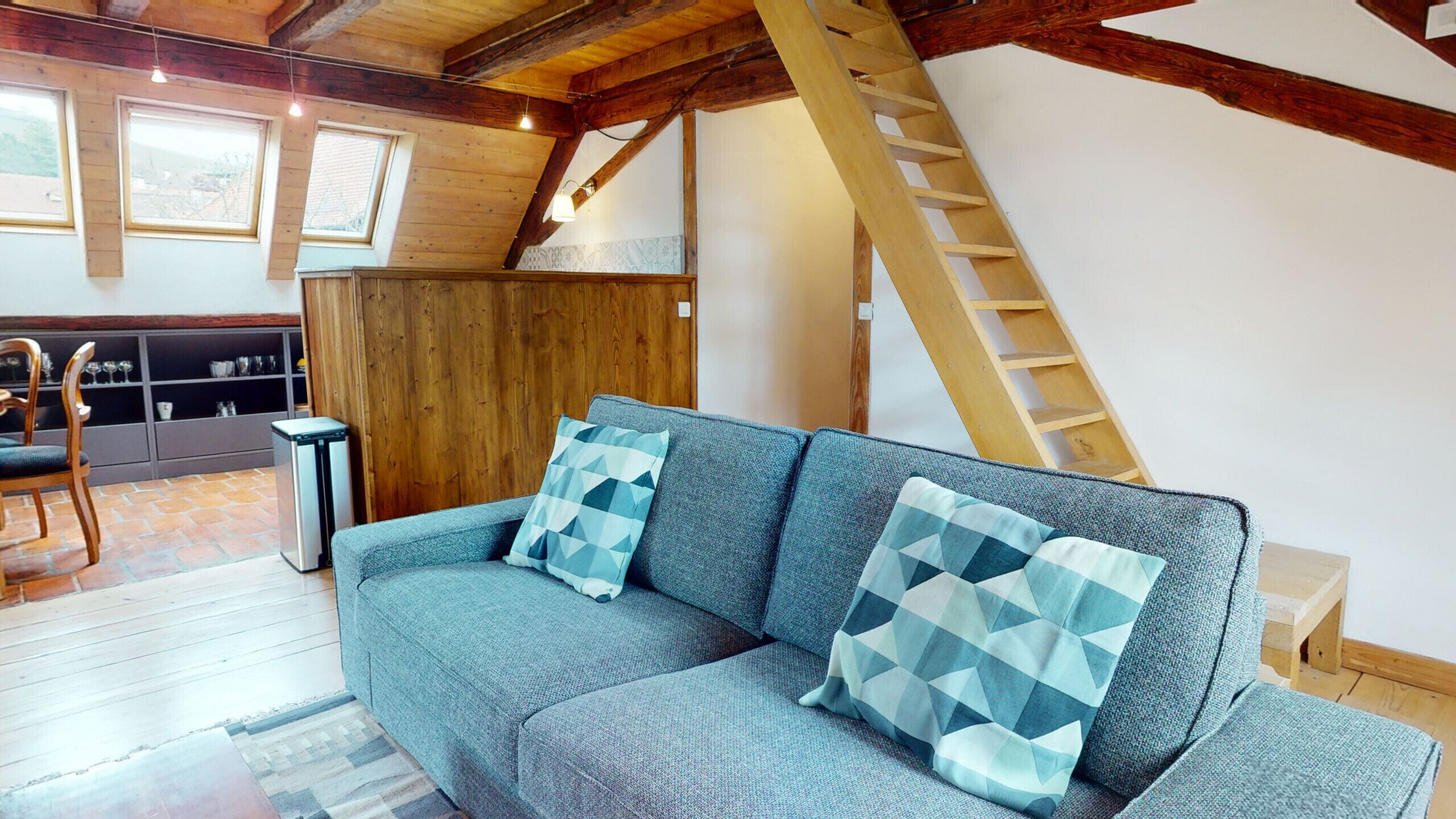 Ein Loft-Wohnzimmer mit einer Couch, gemusterten Kissen, einer Holzleiter und Blick ins Freie durch ein Fenster.
