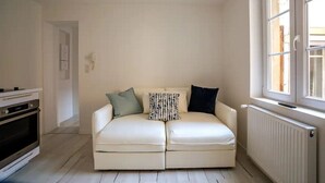 Un espai vital reduït amb un sofà, un radiador, una finestra i una cuina.