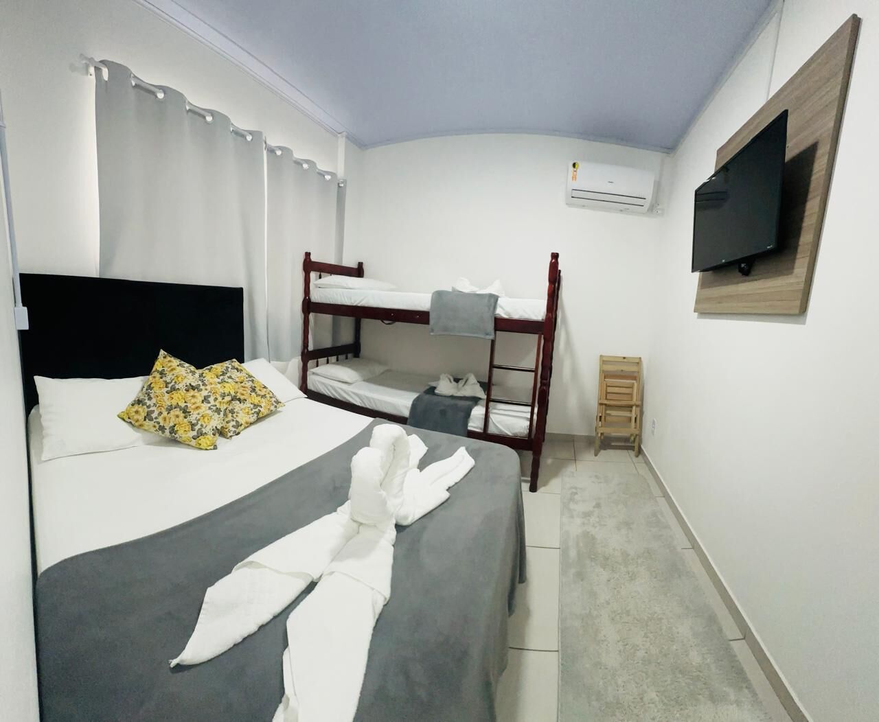 A hotel room with a bunk bed, a single bed, a flat-screen TV mounted on the wall, and an air conditioning unit.