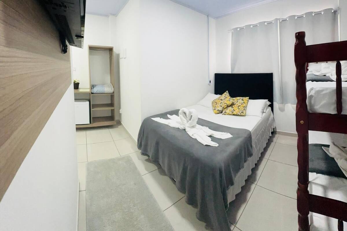 Quarto de hotel com uma cama, cabeceira de madeira, guarda-roupa e uma pequena mesa com uma cadeira.