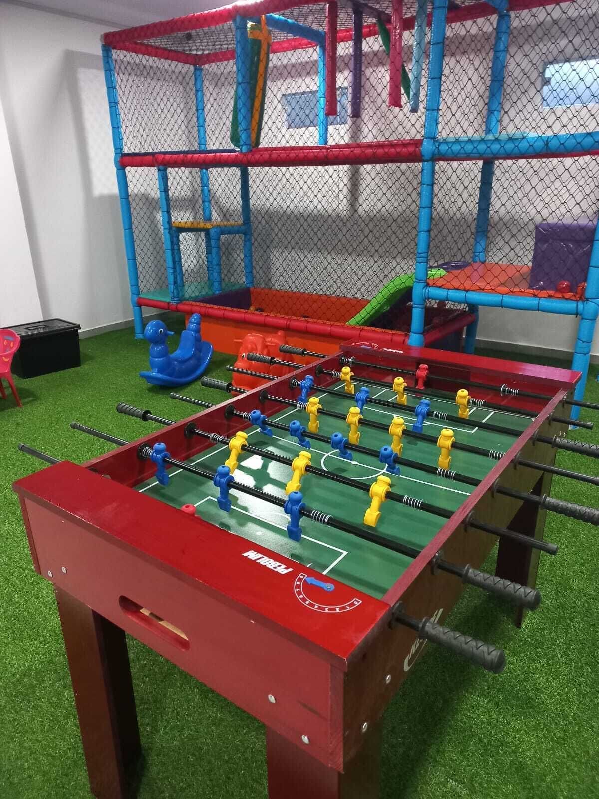 A foosball table with a red and green playing surface, surrounded by a blue and red playpen.