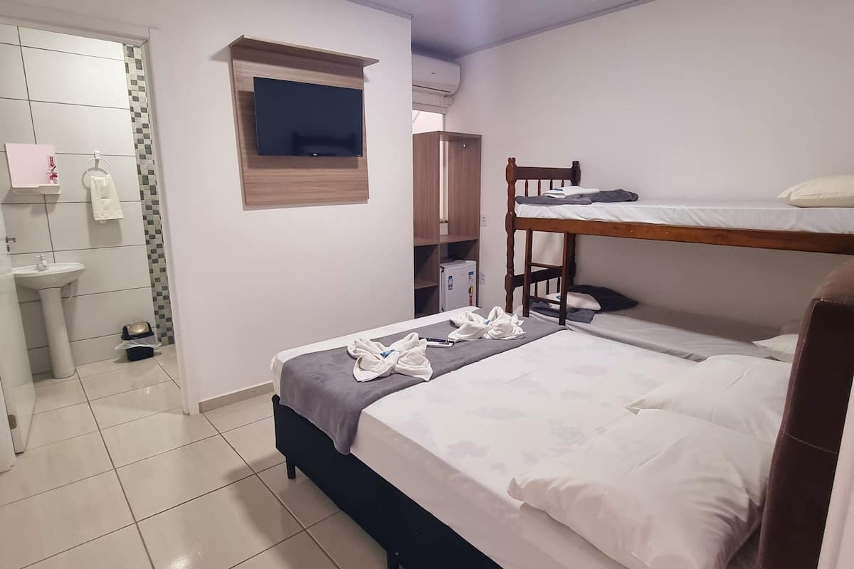 Quarto de hotel com cama beliche, mesa de cabeceira, espelho, armário pequeno e televisão presa à parede.