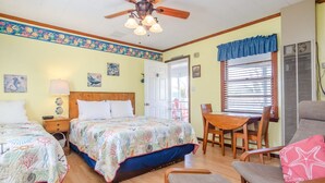 Une chambre avec deux lits, un ventilateur de plafond, une table avec des chaises et une fenêtre avec des stores.