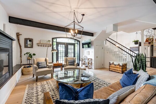 A spacious living room with a round glass coffee table, a sofa with blue cushions, a fireplace, and a staircase leading to an upper level.