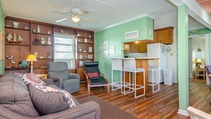 Un salon confortable avec un canapé, un fauteuil et une petite table. On y trouve une bibliothèque remplie de livres et d’objets décoratifs, ainsi qu’un coin cuisine avec un réfrigérateur et des placards.