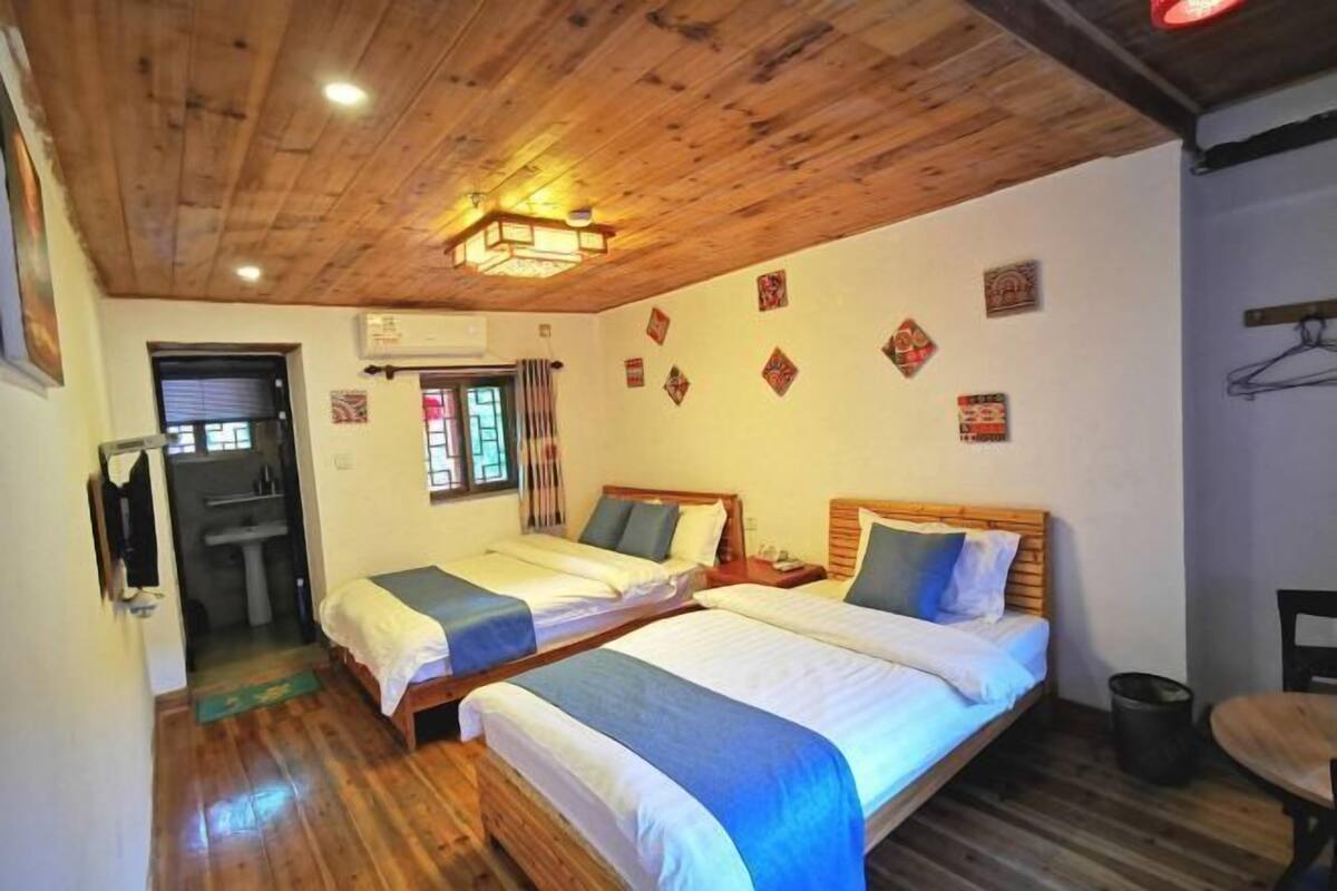 A room with two beds, wooden flooring, a ceiling light fixture, a window with curtains, and a wall-mounted air conditioning unit.