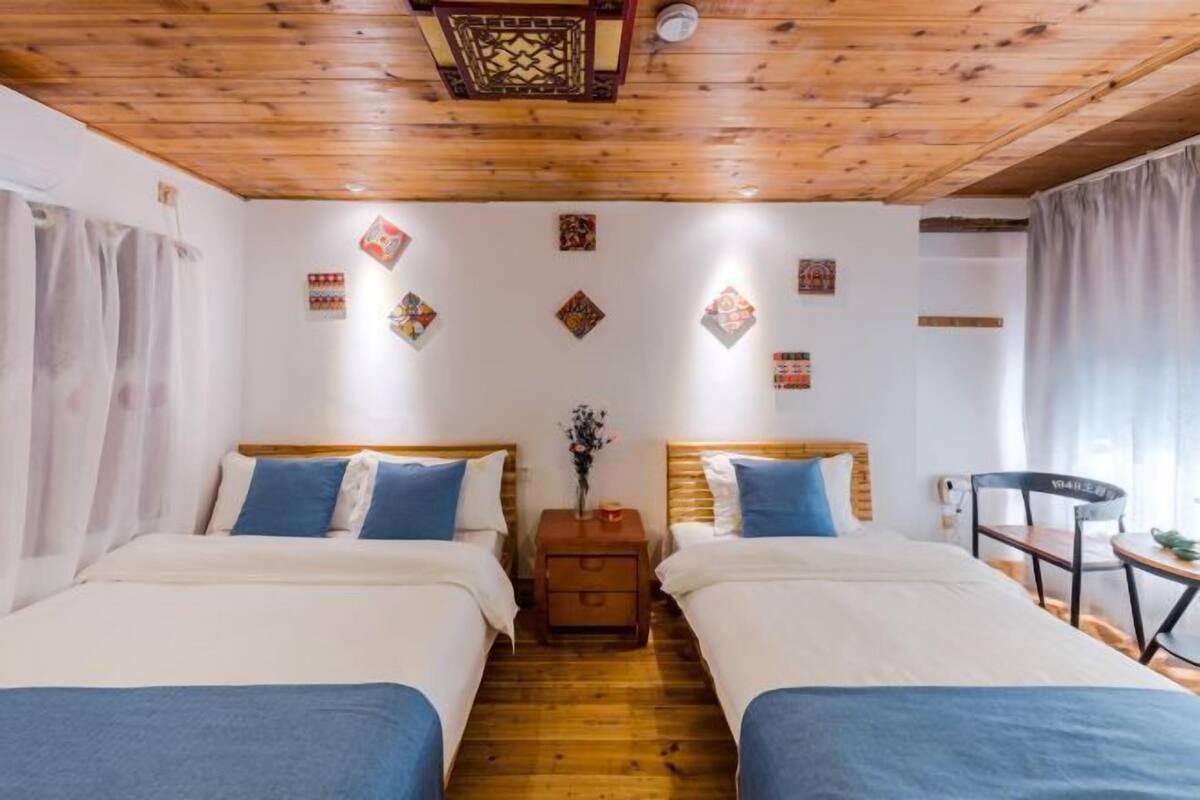 A room with two beds, wooden flooring, a ceiling fan, and a small table with a chair.