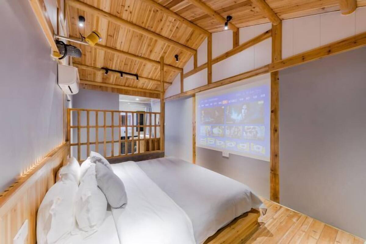 A room with a wooden ceiling, a bed with white bedding, a projector screen, and a wooden railing.