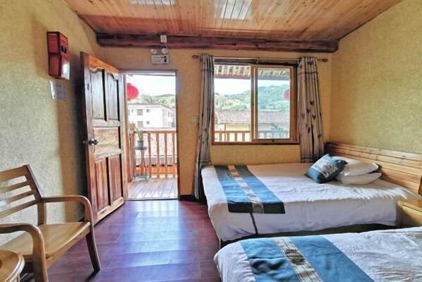 A room with two beds, a wooden chair, and a balcony with a view of buildings and hills.