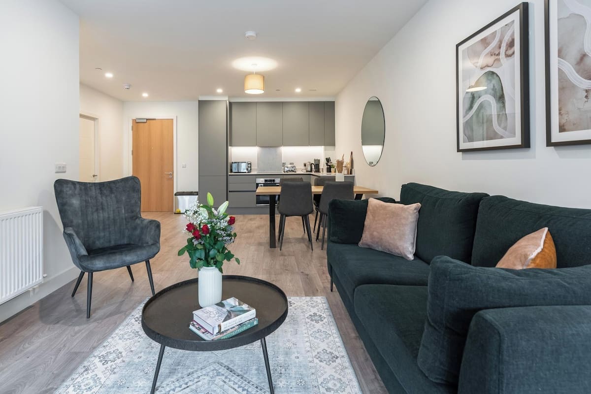 A modern living room with a dark green sofa, a grey armchair, and a coffee table with books and flowers. A kitchen with grey cabinets and a dining area with a table and chairs is visible in the background.
