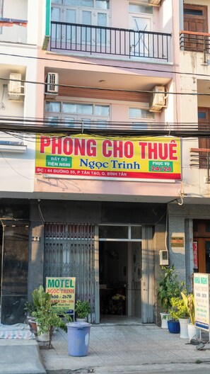 Un edificio con letreros que indican 'Phong Cho Thu' y 'Ngoc Trinh', señalando servicio disponible las 24 horas. Se observa una entrada con una cancel y un letrero de motel que muestra su horario de atención.