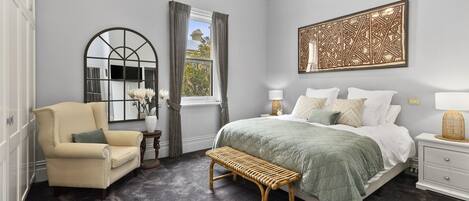 A bedroom with a bed, a chair, a bench, a nightstand, a lamp, a mirror, and a wall art piece.