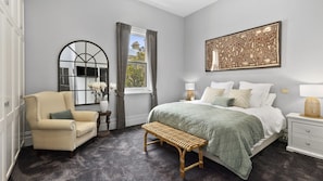 A bedroom with a bed, a chair, a bench, a nightstand, a lamp, a mirror, and a wall art piece.
