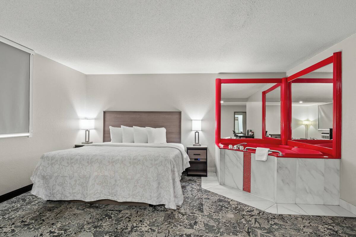 A hotel room with a bed, bedside tables, a bathroom with a red sink and mirror, and a patterned carpet.