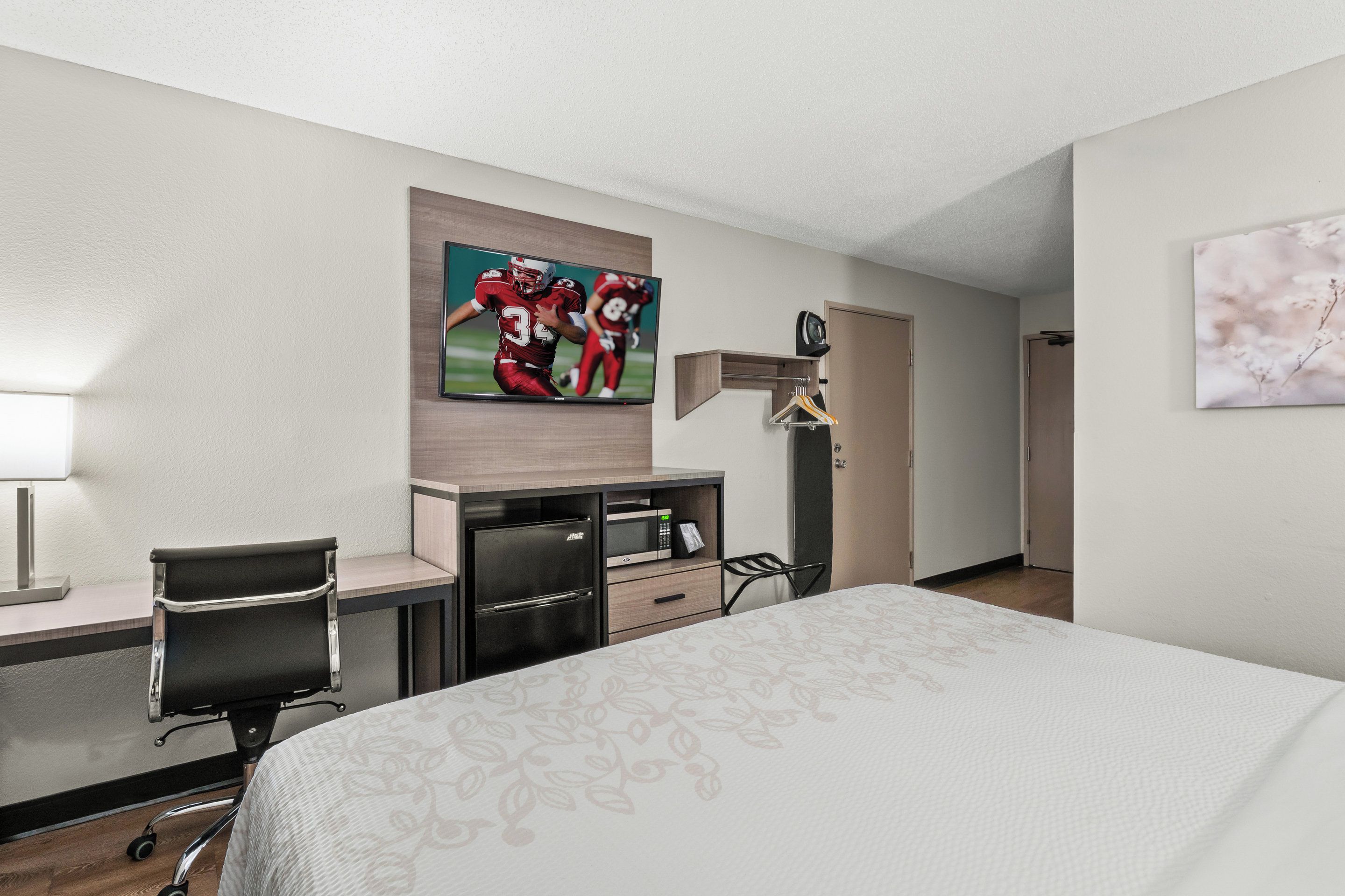 A hotel room with a bed, a desk with a chair, a TV mounted on the wall, and a door leading to another room.