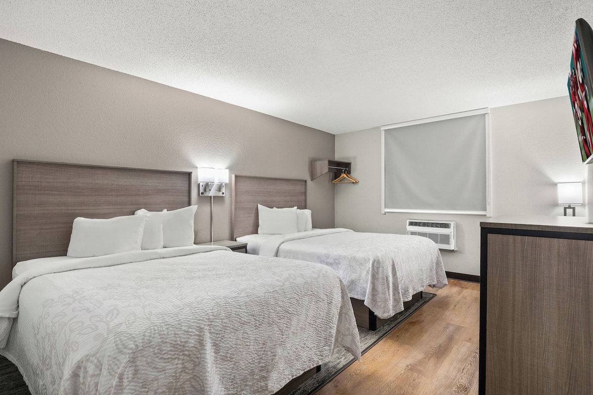 A hotel room with two beds, a desk, a wardrobe, and a large window.